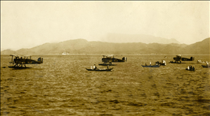 Three single propellor planes resting on pontoons in a large bay surrounded by small canoe like boats. In the distance a large ship and mountains. 